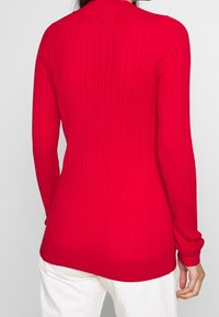 Pull rouge en maille avec texture côtelée, manches longues et encolure arrondie. Présente une silhouette cintrée et un design uni.