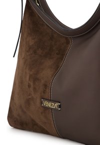 Brown suede and leather handbag featuring smooth and textured panels, contrasting stitching, and a gold logo accent on the front.