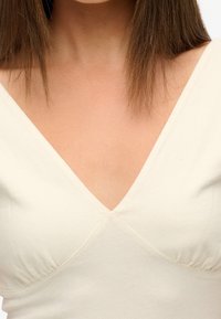 Cremefarvet top med V-udskæring og drapering ved barmen, lavet af blødt, glat stof og designet til en tætsiddende pasform.