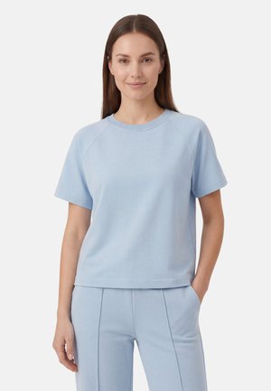 Kobieta z prostymi brązowymi włosami, ubrana w jasnoniebieską bluzkę z krótkim rękawem i pasujące spodnie, jedna ręka w kieszeni, neutralne tło.