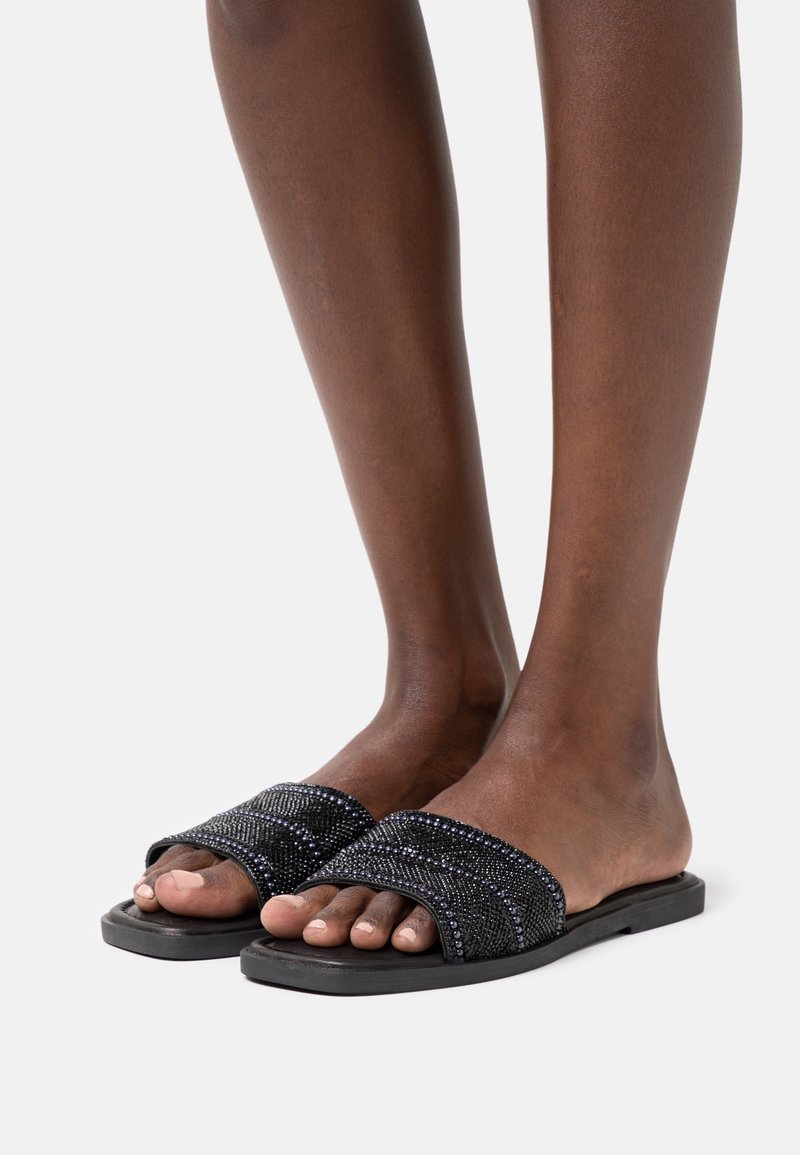 Pieds à la peau foncée portant des sandales slides noires avec des larges brides texturées et perlées sur un fond clair uni.