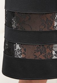 Mustan hameen, jossa on vaakasuoria verkko-osioita kukkakuvioisella pitsillä, korostettuna tummilla mustilla nauhoilla. Pehmeä rakenne ja jämäkkä muotoilu.