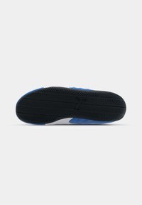 Semelle en caoutchouc noir avec motif de bande de roulement semblable à un pneu sur une chaussure de sport bleue et blanche, vue par le dessous sur fond blanc.
