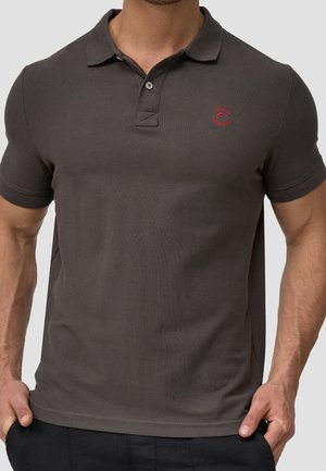 Homme portant un polo gris foncé ajusté avec un logo rouge brodé sur la poitrine gauche, debout les mains dans les poches.