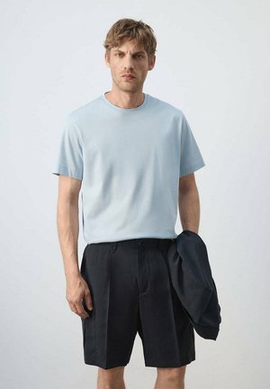 Homme portant une chemise à manches courtes bleu clair et un short noir tenant une veste foncée sur un fond clair uni.