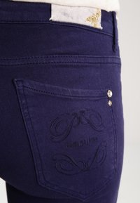Dark purple denim jeans featuring a back pocket with embroidered logo and two decorative silver studs. Smooth texture with a fitted shape.