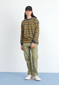 Long-sleeve striped sweater in green, orange, and gray, with ribbed cuffs. Paired with light green pants and white sneakers. Black cap worn.