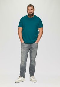 Turkusowa koszulka, krótkie rękawy, luźny krój; jasnoszare dżinsy; białe sneakersy z beżowymi akcentami; casualowy zestaw na stojącym modelu mężczyzny.