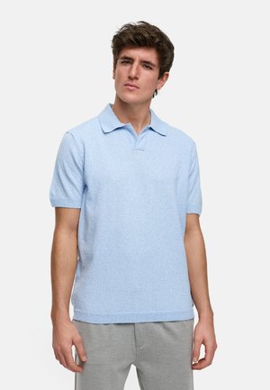 Polo en maille texturée de couleur bleu clair à manches courtes, avec un col classique et un ourlet droit, associé à un pantalon gris.
