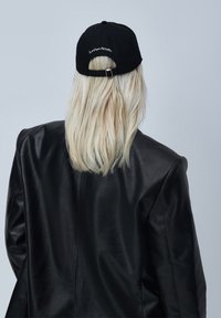 Svart baseballcaps med bøyd skygge, justerbar bakre stropp og hvit brodering av "Le Ger Studio". Båret med en svart skinnjakke.