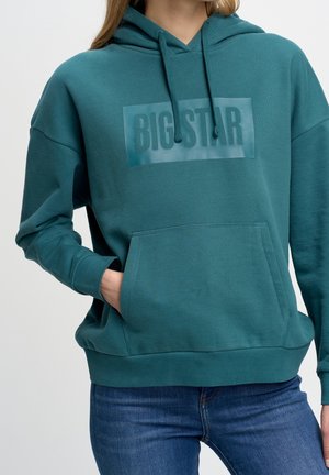 Türkisfarbener Hoodie mit Fronttasche und dem Schriftzug "BIG STAR", getragen von einer Person mit der Hand in der Tasche und blauer Jeans.