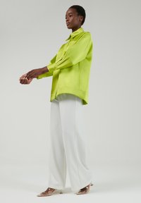 Chaqueta verde lima con cuello abotonado, mangas largas y un dobladillo ligeramente curvado, combinada con pantalones anchos blancos y tacones transparentes.