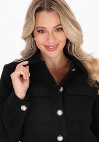 Črna jakna z gumbi, izdelana iz teksturirane tkanine, z oblogo iz srebrne barve, dvema sprednjima žepoma in ovratnikom. Model drži ovratnik.