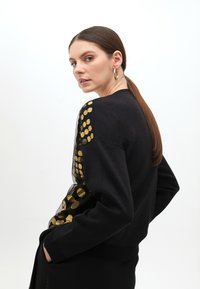 Black sweater featuring gold polka dot accents on one shoulder. The texture is smooth with a fitted design and ribbed hem.