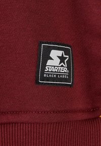 Burgunderfarbener Sweatstoff mit geripptem Saum, versehen mit einem aufgenähten Logo-Patch in Schwarz und Weiß von Starter Black Label auf der Vorderseite.