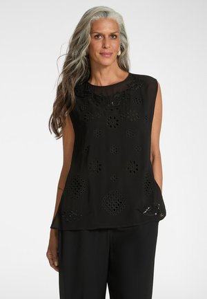 Femme aux longs cheveux gris portant un haut noir sans manches avec des découpes florales et un pantalon noir, debout devant un fond uni.