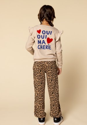 Kind met gevlochten haar draagt een beige sweatshirt met ruches waarop "Oui Oui Ma Cherie" staat, versierd met rode harten, een panterprint broek en zwarte Adidas-schoenen.