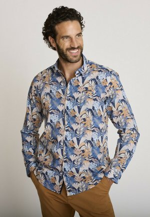 Homme aux cheveux bouclés et barbe souriant, portant une chemise à manches longues bleu et marron à motif feuille et un pantalon marron, les mains dans les poches.