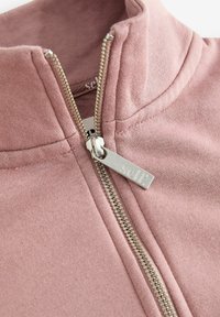 Felpa zip-up rosa chiaro con colletto alto, zip metallica con etichetta che riporta "self." e un tessuto morbido e strutturato.