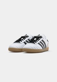Par de sapatilhas Adidas Samba brancas com riscas pretas, atacadores pretos e solas de borracha caramelizada, colocadas sobre uma superfície branca.