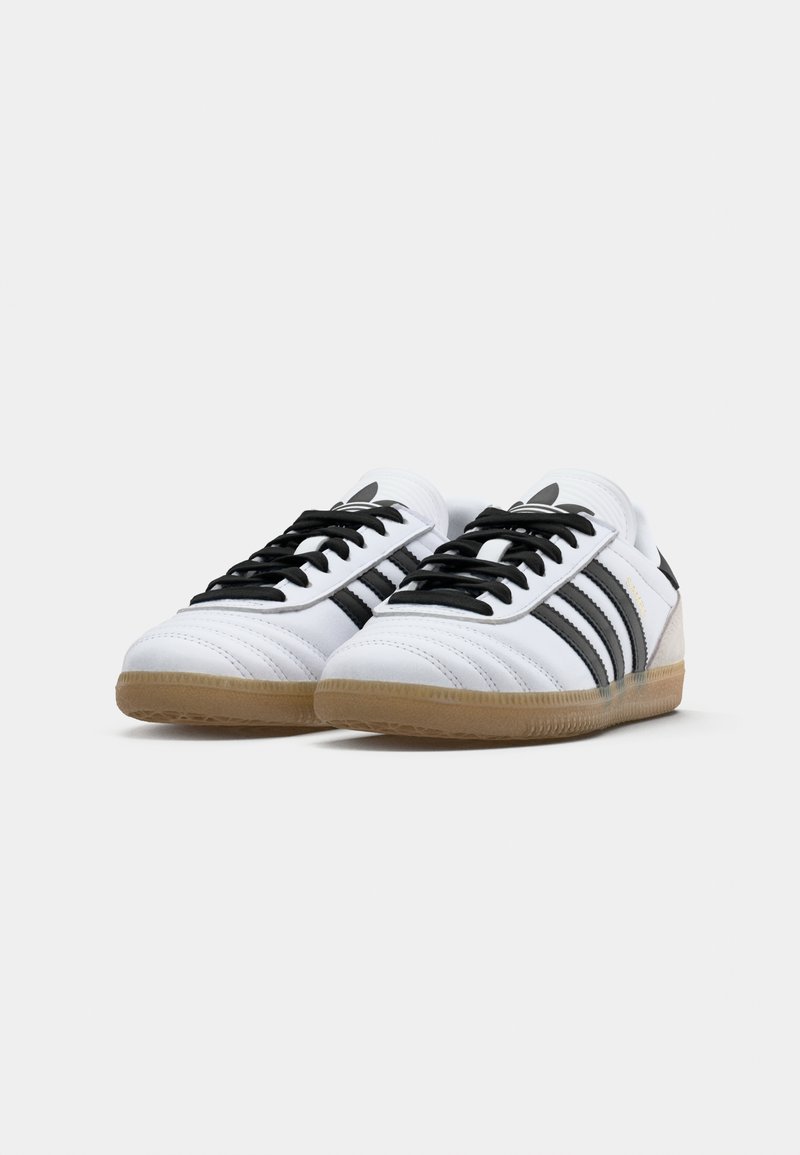 Ein Paar weiße Adidas Samba Sneaker mit schwarzen Streifen, schwarzen Schnürsenkeln und Sohlen aus braunem Gummi auf einer weißen Fläche platziert.