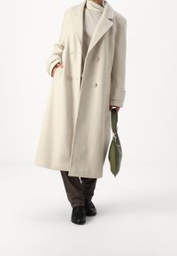 Cappotto in lana beige chiaro con design doppiopetto, ampi rever e vestibilità oversize. Abbinato a pantaloni marrone scuro e una borsa verde.