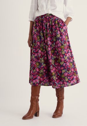 Mujer que lleva una blusa blanca, una falda midi floral colorida y botas de cuero marrón hasta la rodilla con tacón, de pie con las manos en los bolsillos.