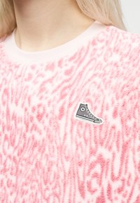 Ροζ χνουδωτό πουλόβερ με διακριτικό μοτίβο ζωολογικού τύπου και μικρό μαύρο και λευκό αυτοκόλλητο παπουτσιού στο στήθος.