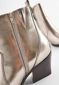 Botas de tobillo plateadas metálicas con un acabado texturizado, doble cremallera y un tacón bloqueado con ranuras horizontales para mayor agarre.