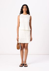 Robe tricotée blanche en deux pièces avec un haut sans manches et une jupe présentant des motifs texturés. Modèle tenant un sac à main marron, portant des tongs noires.