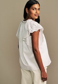 Blusa bianca con maniche a ruffle in pizzo ad occhiello intricati, scollatura arricciata e tessuto leggermente testurizzato. Abbinata a pantaloni di colore chiaro.