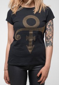 Black t-shirt featuring a large gold rhinestone Prince symbol in the center. Short sleeves, crew neck, and soft fabric. Fitted style.