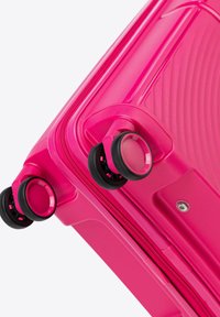 Valigia rigida rosa con superficie testurizzata, dotata di doppie ruote nere e chiusura con zip. Gli accenti includono un design di ruote circolari.