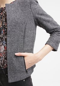 Blazer grigio textured con un design cropped, dotato di una tasca con zip visibile e maniche a tre quarti, indossato sopra una blusa floreale.