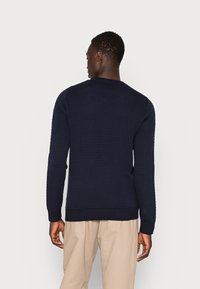 Marineblå strikkegenser med et strukturert mønster, ribbestrikkede mansjetter og kant, med rund hals. Brukt over lysebeige bukser.