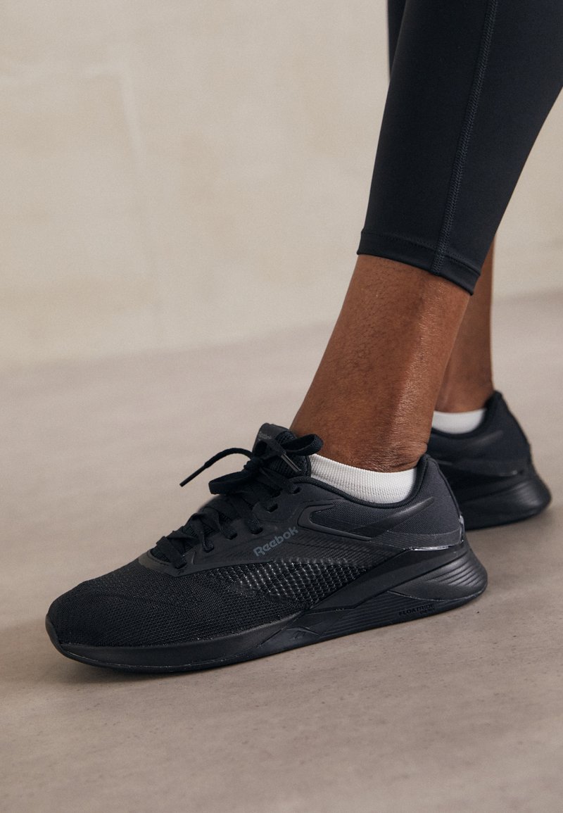 Zapatillas deportivas negras con parte superior de malla y sintético, puntera redonda, cordones y cuello acolchado; combinadas con calcetines blancos hasta el tobillo y leggings negros.