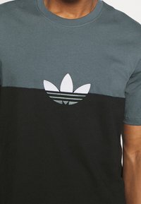 Camiseta de algodón de manga corta con la parte superior en color azul verdoso, la parte inferior en negro y un gran logo bordado de Adidas en blanco y azul verdoso.