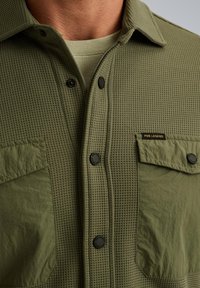 Olijfgroene shirt met wafelstructuur, knoopsluiting, twee borstzakken en zwarte knopen. Opvallend logo-label op de zak.