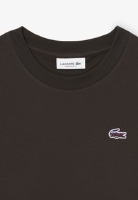 T-shirt nera oversize con scollatura tonda. Presenta un logo del coccodrillo Lacoste ricamato in viola, contornato di bianco.