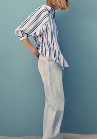 Persona che indossa una camicia a strisce blu e bianche a maniche lunghe e jeans azzurri chiari, in piedi di lato contro un muro blu uniforme.