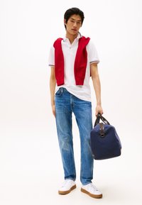 Νεαρός άνδρας που φοράει λευκή πόλο μπλούζα και μπλε τζιν, με κόκκινο πουλόβερ περασμένο στους ώμους, κρατώντας μια βαλίτσα τύπου duffel σε ναυτικό μπλε χρώμα.