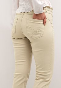 Beige skræddersyede bukser med en glat tekstur, der har buede baglommer og subtile syningsdetaljer. Nærbillede af bagsiden.