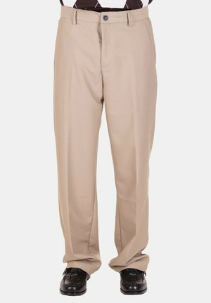 Pantaloni beige a gamba dritta indossati con scarpe in pelle intrecciata nere su una persona in piedi davanti a uno sfondo semplice.