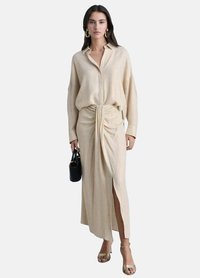 Beige lange jurk met een geknoopte taille, een blouse met kraag en knopen, en een zijsplit. Model houdt een kleine zwarte tas vast en draagt sandalen met banden.