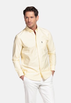 Mann trägt blassgelbes Hemd mit Brusttasche und weiße Hose, steht mit den Händen in den Taschen und blickt zur Seite.