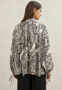 Sort og hvid mønstret bluse med lange ærmer, rynkede manchetter, bindebånd ved nakken og et løst, flydende design.