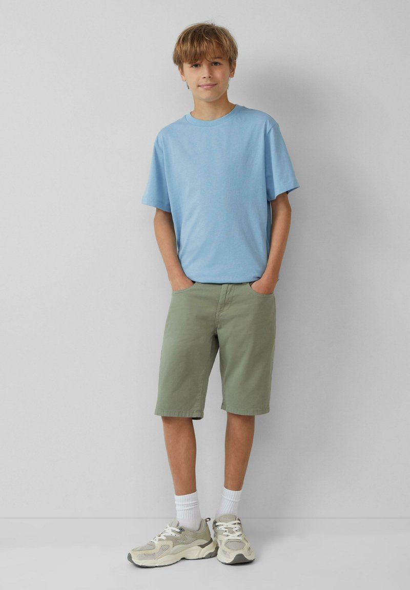 Lichtblauw t-shirt met korte mouwen, groene shorts, witte enkelsokken en beige sneakers met mesh en accentdetails. Staand met de handen in de zakken.