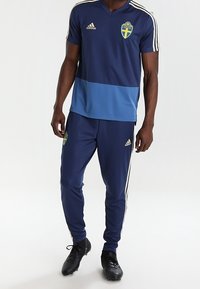 Mörkblå Adidas träningsdräkt med en blå rand, svensk klinga, guldaccenter och tre vita ränder på axlarna och sidorna av byxorna.