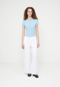 Ανοιχτό γαλάζιο εφαρμοστό μπλουζάκι με ριμπ ύφανση και κοντά μανίκια, με λεπτομέρεια κουμπιών στο λαιμό· συνδυασμένο με λευκό παντελόνι φαρδιάς γραμμής. Μαύρα παπούτσια slip-on ολοκληρώνουν την εμφάνιση.