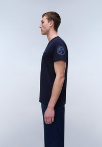 Young man standing in profile wearing a dark blue t-shirt with a circular patch on the sleeve and matching dark pants.